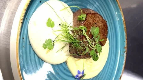 The cook's hands scroll through a blue plate with pork steak and white sauce Stock Footage 246474364