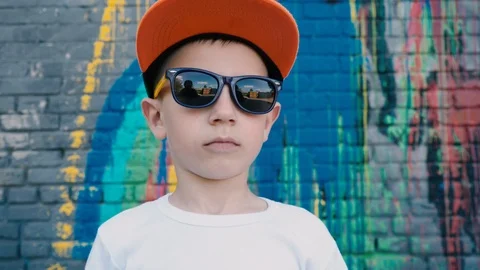 A cool 5 year old boy in a cap and sunglasses posing on the camera near the wall Video stock 89688390