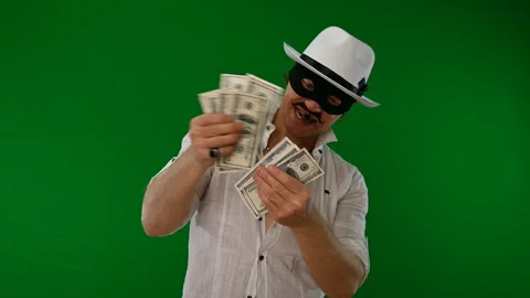 Cool bad guy with a black mask, white hat, cigar and money on a green backdrop 스톡 동영상 141856166