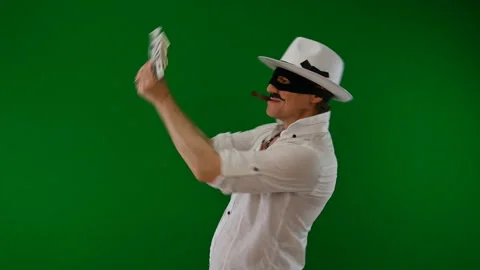 Cool bad guy with a black mask, white hat, cigar and money on a green backdrop Stock Footage 141856390