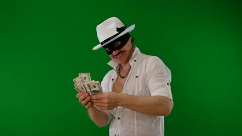 Cool bad guy with a black mask, white hat, cigar and money on a green backdrop Stock Footage 141856409