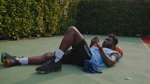 Cool black guy is resting after basketball training, lying on ground and using Stock Footage 227364561