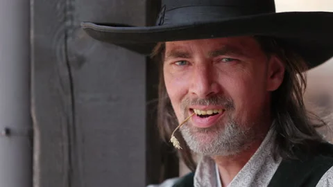 A cool cowboy with a grey beard chews on a blade of grass.He scowls at the Stock Footage 163039529
