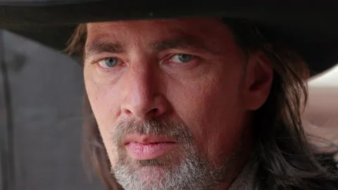 A cool cowboy with a grey beard chews on a blade of grass.He scowls at the Stock Footage 163040122