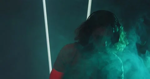 Cool deejay young long haired guy wearing headphones in smoke on party in Stock Footage 158044294