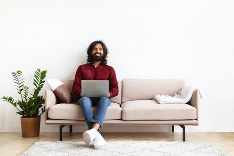 Cool indian guy chilling on couch at home, using laptop 写真素材