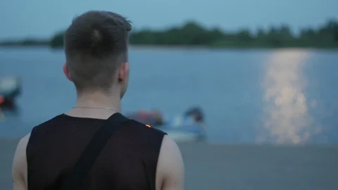 Cool young guy walking to the water on the embankment in the dusk, Slow motion Video stock 113066127