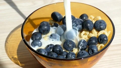Cornflakes rings and blueberries into empty glass bowl pour milk, slow motion Stock Footage 96977095