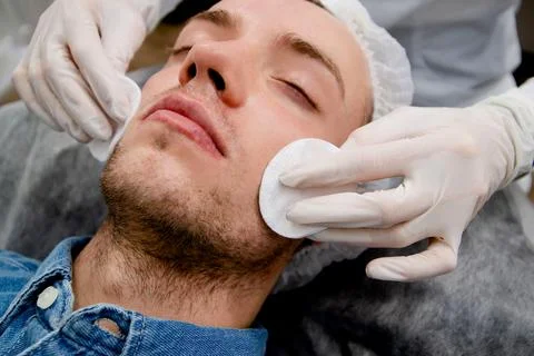 Cosmetologist is cleaning man's face using cleanser and pads to remove acne and Stock Photos