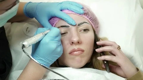 Cosmetologist is making an eyebrow permanent makeup while woman talking on the Stock Footage 74215128