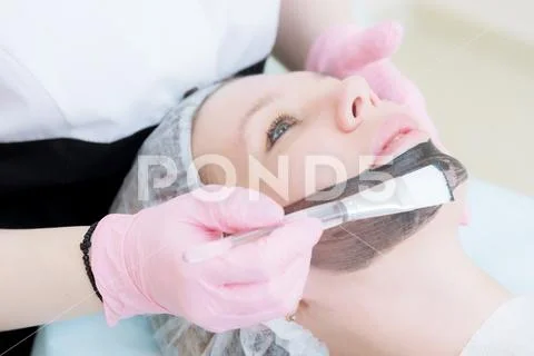 The cosmetologist in pink gloves with a brush applies a carbon mask for peeling Stock Photos