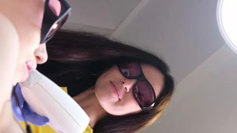 A cosmetologist in protective glasses performs laser hair removal on a young Stock-Footage 250356644
