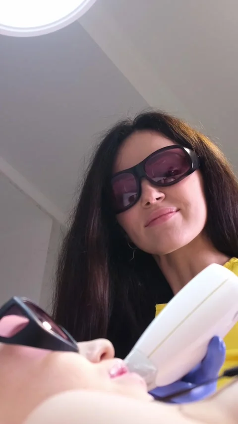A cosmetologist in protective glasses performs laser hair removal on a young Stock Footage 250384899