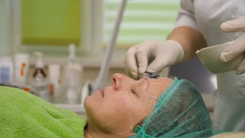 Cosmetologist puts gel on face of elder woman before face lifting procedure at Stock Footage 102400530