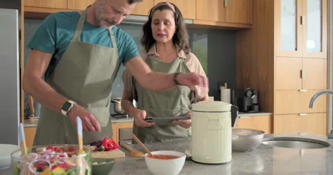 Couple in aprons using tablet in kitchen chopping on cutting board opening Stock Footage 341242062