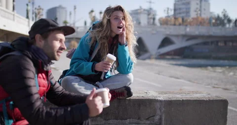 Couple of Backpackers Rest by the River in the City, Drinking Coffee and Stock Footage 344830233