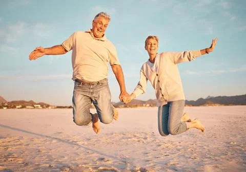 Couple, beach and active seniors hold hands while jumping in sand, happy and Stock Photos