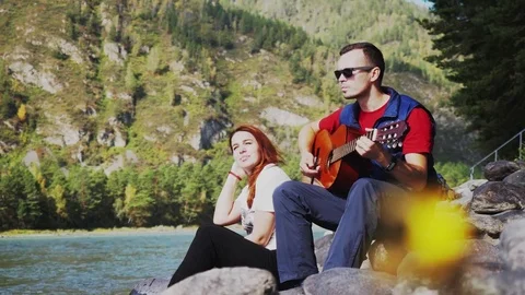 Couple on the beach playing guitar on a summer day next to mountain river Stock Footage 80370868