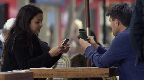 Couple both checking their iPhones, sitting outside at a cafe table Stock Footage 65331721