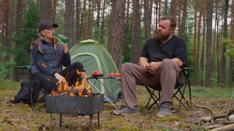 Couple camping in forest drinking beer and talking by campfire Stock Footage 327134901
