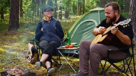 Couple camping in forest with man playing guitar Stock Footage 327136677