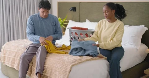 Couple carrying laundry basket onto bed, sitting, sorting and folding tops and Stock Footage 346614988