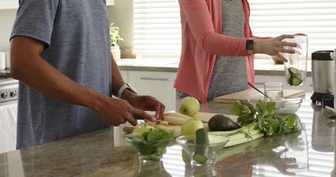 Couple chopping bananas and adding cucumber to blender cup at kitchen island, Stock Footage 344534959