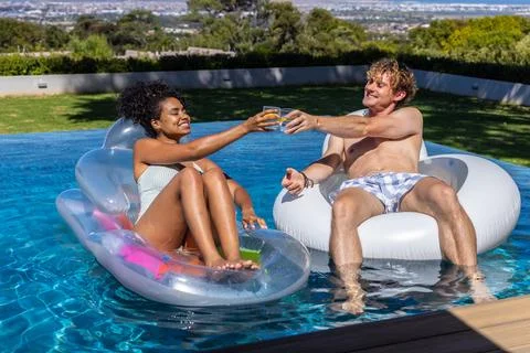 Couple clinking glasses while floating on lounger and donut in backyard pool Foto stock