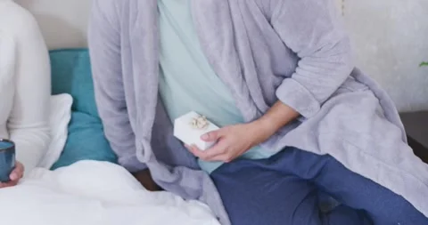 Couple on cozy bed, man in gray robe placing gift box, offering to woman to Stock Footage 344402297