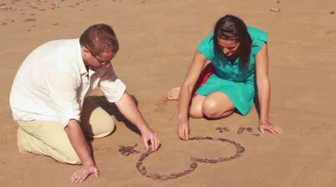 Couple creating heart shape with stones in the sand and kissing Stockbeeldmateriaal 10892196