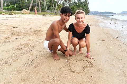 Couple crouching on the beach while drawing a heart shape on the sand Stock Photos