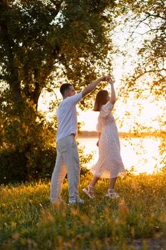 Couple dancing joyfully in a sun-drenched meadow by the water at sunset Stock Photos
