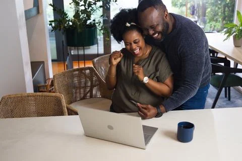 Couple embracing and celebrating while using silver laptop at dining table with Stock Photos