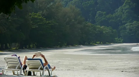 Couple on empty beach, relaxed, reading on sunbeds in a tropical resort Stock Footage 60259818