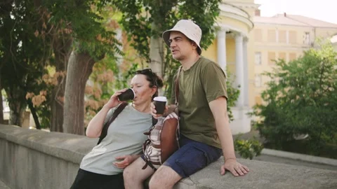 Couple enjoying beverages while resting on a city wall under the shade of trees Stock Footage 301099492
