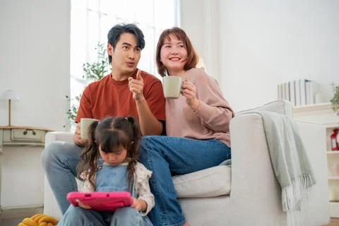 Couple enjoys drinks on a sofa while child plays with a tablet in a bright .. Foto stock