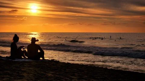 Couple enjoys sunset on the beach while surfers ride waves in the background Stock Photos