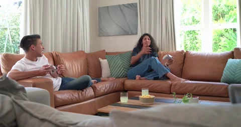 Couple gaming on tan couch with snacks, laughing and holding controllers after Stock Footage 341468113