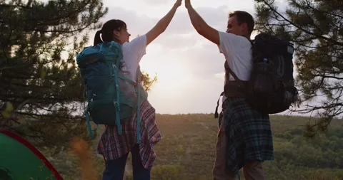 Couple hikers high-five at forest camp with backpacks, celebrating sunset Stock Footage 329910198