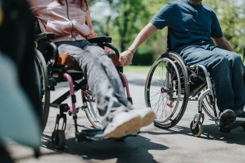 A couple holding hands while sitting in wheelchairs outdoors on a sunny day Foto stock