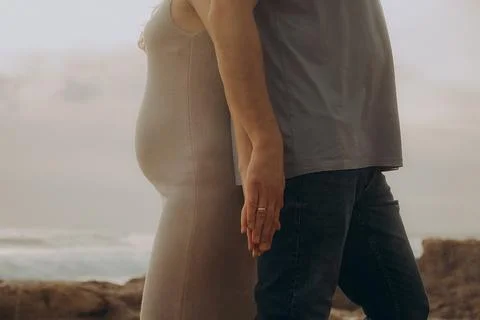 Couple holding hands while standing on rocky shore at sunset near ocean, clou Stock Photos