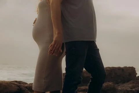 Couple holding hands while standing on rocky shore at sunset near ocean, clou Stock Photos