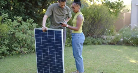 Couple holding solar panel on lawn, woman tapping phone, pointing toward roof Stock Footage 340974707