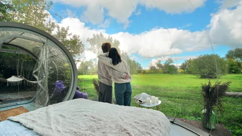 A couple hugging each other inside a bubble tent at glamping. Lush forest around Stock Footage 218480215