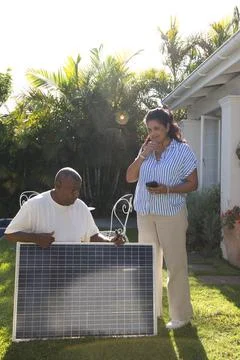 Couple inspecting solar panel on lawn, man in white tee holding tool, woman Fotos Stock