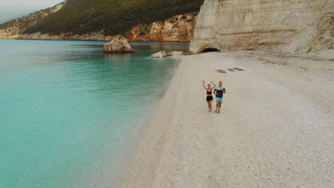 Couple joyfully running along tranquil turquoise sea with pebble beach and rocky Stock Footage 342366590