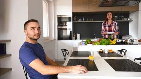 Couple in the kitchen. Man is sitting behind the table. Girlfriend brings food Video stock 61856191