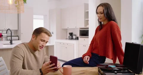 Couple at kitchen table, man turning phone toward woman, showing red smartphone Stock Footage 342608157