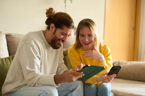 Couple laughing and using tablet and smartphone on sofa at home Stock Photos