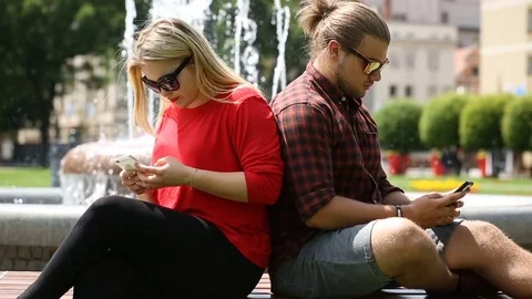 Couple leaning on each other next to the fountain and texting on smartphones Stock Footage 77484123